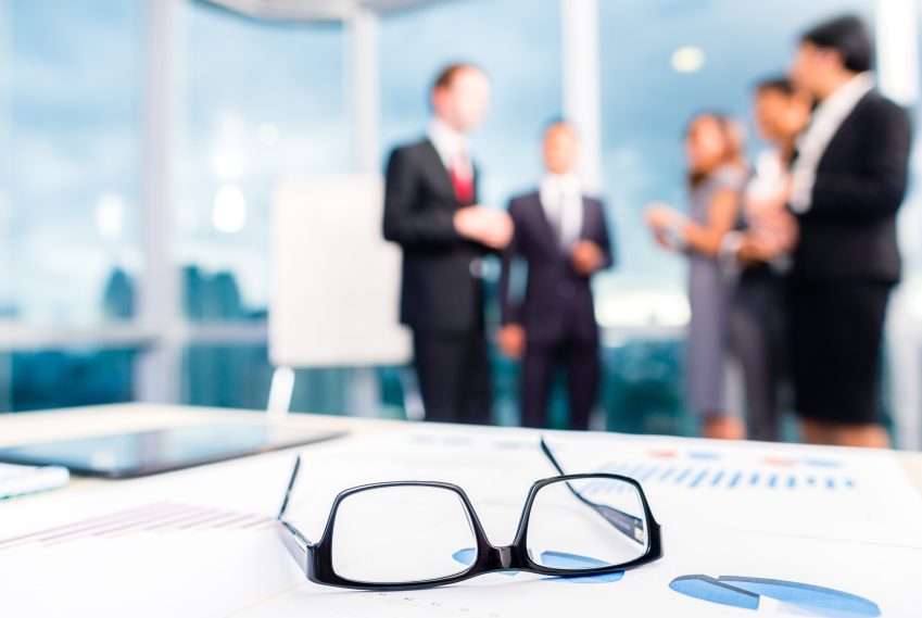 Group of business people standing in an office with a pair of glasses on a table.